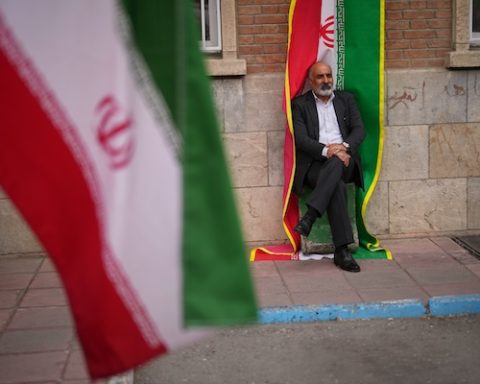 A man sits beside an Iranian flag banner during a government-sponsored protest attended by medical workers against the U.S.-Israeli military campaign outside Imam Khomeini Hospital in Tehran, Iran, Monday, April 6, 2026. (AP Photo/Francisco Seco)