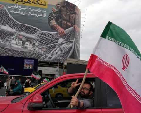 A man flashes a victory sign as he carries an Iranian flag in front of an anti-U.S. billboard depicting the American aircrafts into the Iranian armed forces fishing net with signs that read in Farsi: "The Strait of Hormuz will remain closed, The entire Persian Gulf is our hunting ground," at the Eqelab-e-Eslami, or Islamic Revolution Square in downtown Tehran, Iran, Monday, April 13, 2026. (AP Photo/Vahid Salemi)