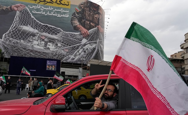 A man flashes a victory sign as he carries an Iranian flag in front of an anti-U.S. billboard depicting the American aircrafts into the Iranian armed forces fishing net with signs that read in Farsi: "The Strait of Hormuz will remain closed, The entire Persian Gulf is our hunting ground," at the Eqelab-e-Eslami, or Islamic Revolution Square in downtown Tehran, Iran, Monday, April 13, 2026. (AP Photo/Vahid Salemi)