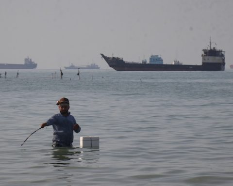 A man stands in the water, appearing to fish, as bulk carriers, cargo ships, and service vessels line the horizon in the Strait of Hormuz off Bandar Abbas, Iran, Monday, April 27, 2026.(Razieh Poudat/ISNA via AP)