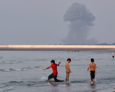 Three boys play in the shallow waters of the Strait of Hormuz, as a plume of smoke rises from an explosion in the background, off Bandar Abbas, Iran, Monday, July 13, 2026. (Razieh Poudat/ISNA via AP)