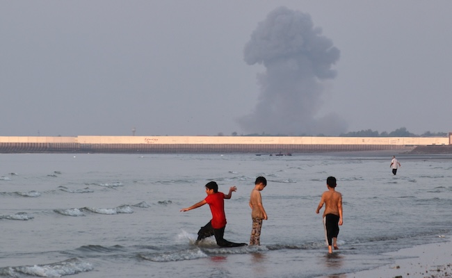 Three boys play in the shallow waters of the Strait of Hormuz, as a plume of smoke rises from an explosion in the background, off Bandar Abbas, Iran, Monday, July 13, 2026. (Razieh Poudat/ISNA via AP)